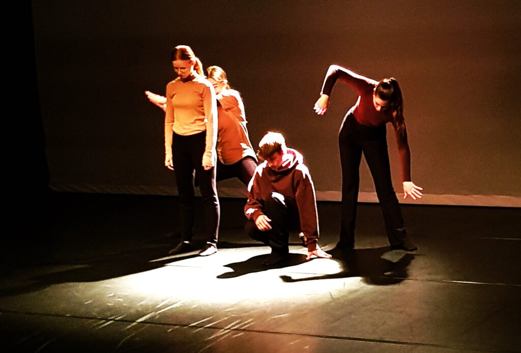 Vier Jugendliche sind in verschiedenen Tanz-Posen auf einer Bühne zu sehen. Sie tragen schwarze Hosen und helle Oberteile. Der Hintergrund ist schwarz, die Jugendlichen sind im Sehweinwerferlicht. 
